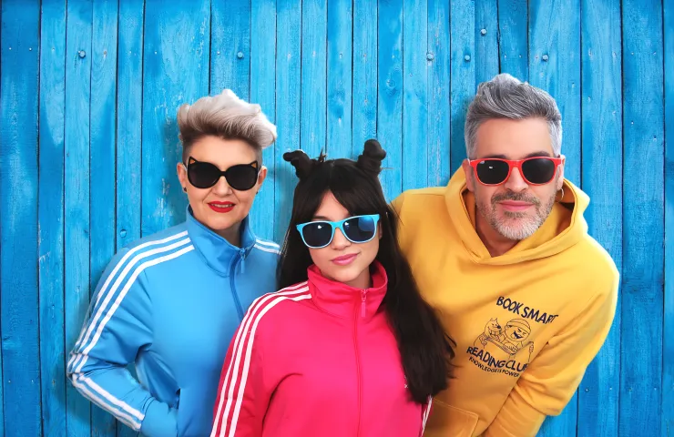 los tres componentes del grupos, gafas de sol de pasta de colores, sudares de chandal de colores, fondo de madera azul