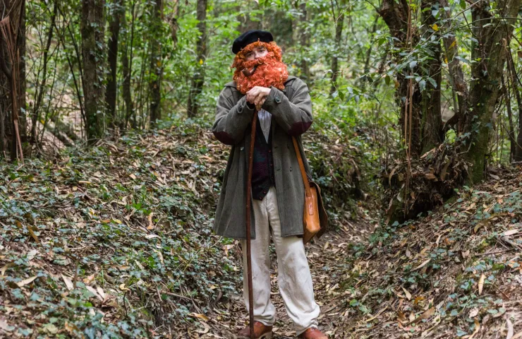 hombre disfrazado con gran barba y pelo pelirrojos, apoya ambas manos en un bastón de paseo, abrigo, boina y zurrón en medio de un bosque