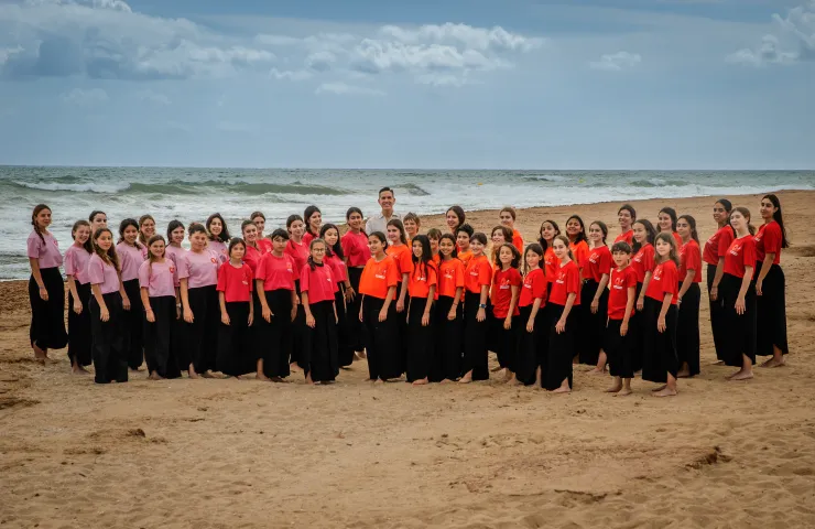 grupo de niñas integrantes del coro en una playa, faldas negras y camisetas rojas o rosas todas agurpados por colores