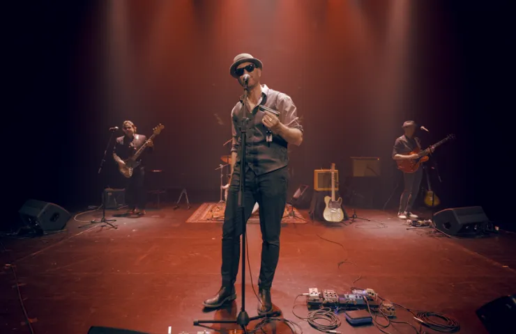hombre con sombrero y gafas de sol cantando en escenario, lleva chaleco y camisa ajustada